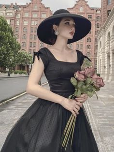 a woman in a black dress and hat holding flowers on the street with buildings in the background