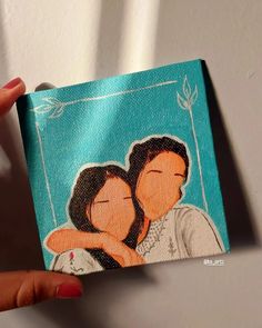 a hand holding up a small painting of two people with their arms around each other