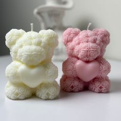 two small teddy bears sitting next to each other on a white counter top, one holding a heart shaped candle