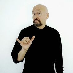 a bald man with a goatee pointing to the side while standing in front of a white wall