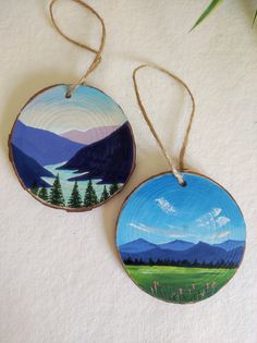 two wooden ornaments with mountains and trees painted on them, hanging from twine strings