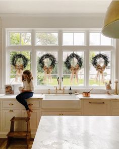 a woman sitting on a kitchen counter in front of a window with wreaths hanging from it