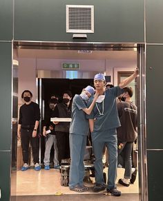 two men in scrubs and masks are standing outside an elevator with their arms around each other