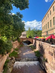 a river running under a bridge next to a tall brick building with red umbrellas