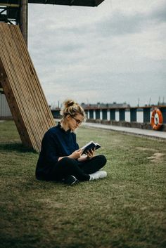 a woman sitting on the ground reading a book in front of a wooden structure with grass