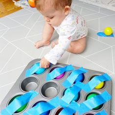 a baby playing with some blue tape on the floor next to muffin tins