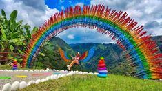 a woman flying through the air next to a rainbow colored sculpture on top of a lush green hillside