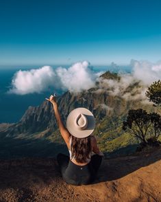 a woman sitting on top of a mountain with her arms in the air and clouds behind her