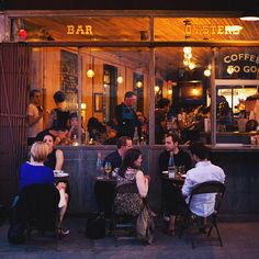 people sitting at tables in front of a bar with lights on the windows and doors