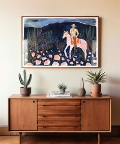 a painting hangs on the wall above a dresser with potted cacti and succulents