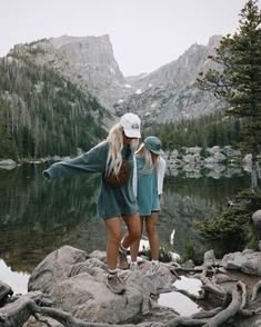 two women standing on rocks looking at the water and mountains in the distance with trees around them