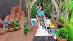 a hand is holding a miniature doll in front of a fake garden area with potted plants