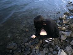 a woman sitting on rocks by the water looking down at her feet in the water