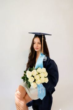 a woman in a graduation cap and gown with flowers sitting on a stool holding a bouquet of white roses