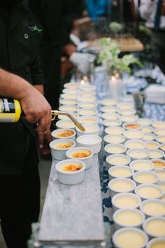 a man pouring sauce into small bowls on a table with other people in the background