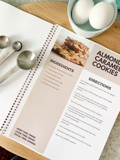 an open recipe book with spoons and eggs in the bowl next to it on a table