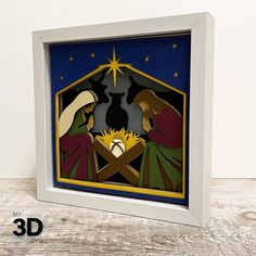 the nativity scene is made out of paper and framed in a white frame on a wooden table