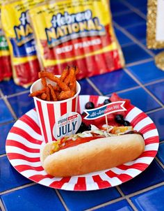 a hot dog on a paper plate with french fries in front of it and two bags of potato chips