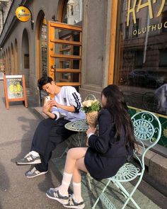 two people sitting on a bench in front of a building eating food and talking to each other