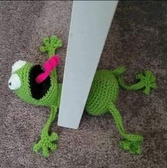 a crocheted green frog with a pink hat on it's head sitting next to a wall