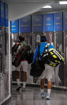 two men with backpacks are walking down the hallway in front of blue and white lockers