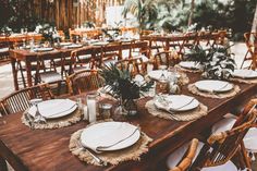 a wooden table topped with white plates and place settings covered in greenery next to tall palm trees