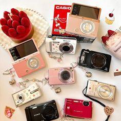 many different types of cameras and accessories on a white surface with a bowl of raspberries in the background