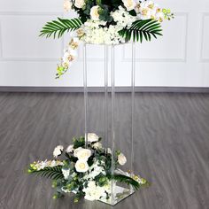 two tall clear vases with white flowers and greenery in them on a wooden floor