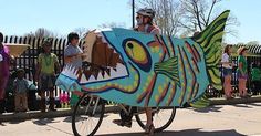 a man riding a bike with a large fish on it's front wheel, while people watch from the other side