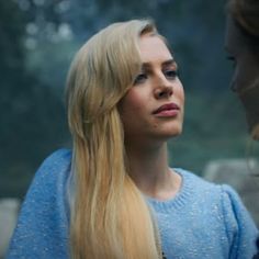 a woman with long blonde hair standing next to another woman in a blue sweater looking at each other