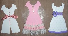 three paper dolls are sitting next to each other on a cloth covered surface, one is wearing a dress and the other has a bow