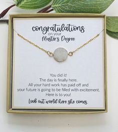 a necklace with congratulations written on it sitting in a box next to a leafy green plant