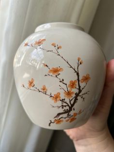 a hand holding a white vase with orange flowers painted on the side and brown branches