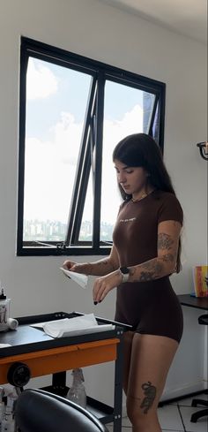 a woman standing in front of a desk with tattoos on her arms and legs, holding a piece of paper