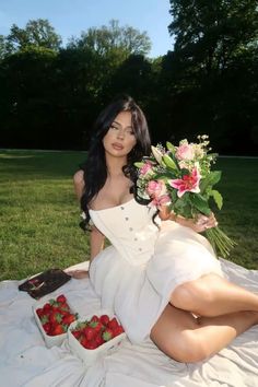 a woman sitting on the ground with flowers and strawberries in her hand while holding a bouquet