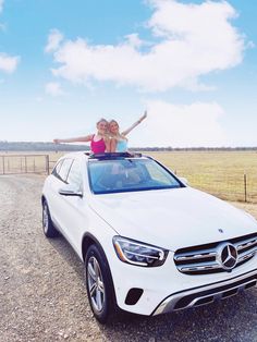 two women standing on top of a white car in the middle of an open field