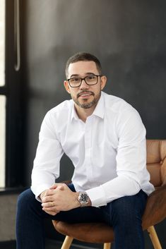 a man sitting in a chair with his hands on his knees, wearing glasses and a white shirt