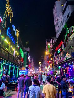 many people are walking around in the city at night with neon lights on buildings and palm trees