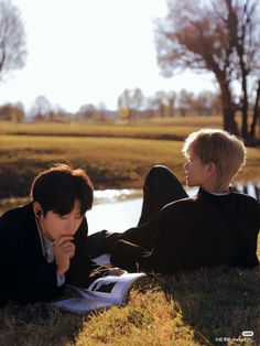 two young men sitting on the grass next to each other, one reading a book