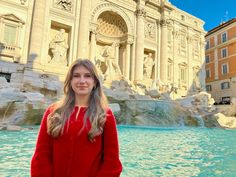 a woman standing in front of a fountain with statues on the sides and buildings behind her