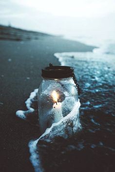 a glass jar with a candle in it sitting on the beach next to the ocean