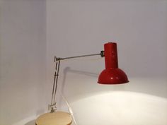 a red table lamp sitting on top of a white wall next to a wooden stool