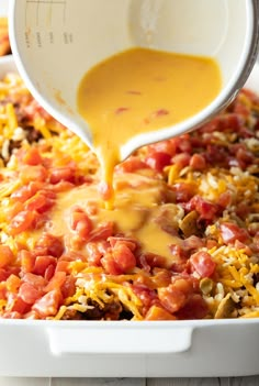 someone pouring cheese on top of a casserole in a white dish with tomatoes and other toppings