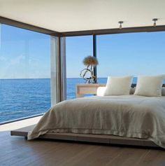 a bed sitting in front of a large window next to the ocean on top of a wooden floor