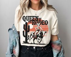 Introducing our "Queen of the Rodeo" graphic tee by Lasso & Lace, perfect for the bold cowgirl who commands the rodeo with style. This Bella Canvas 3001 shirt is soft, lightweight, and comfortable, making it ideal for rodeo days or casual nights out. Featuring a vintage-style graphic of a fierce rodeo queen riding a bull against a sunset backdrop with cacti, this tee captures the essence of Western spirit. Available in white, natural, soft cream, and soft pink, it's perfect for showcasing your l Aesthetic Country, Sunset Backdrop, Cowgirl Gifts, Queen Of, Cowgirl Aesthetic, Western Graphic Tees, Rodeo Shirts, Rodeo Queen, Casual Night Out