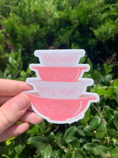 a hand holding three pink and white bowls in front of some bushes with green leaves