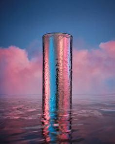 a tall glass sitting in the middle of water with pink and blue clouds behind it