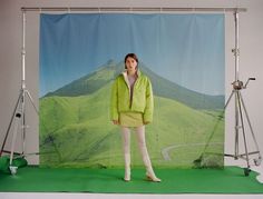 a woman is standing in front of a backdrop with mountains and hills behind her wearing a green jacket