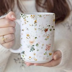 a woman holding a white coffee mug with flowers all over it and gold rings on her fingers