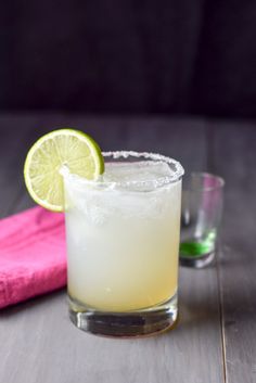 a margarita cocktail with a lime slice on the rim next to a pink napkin and two shot glasses